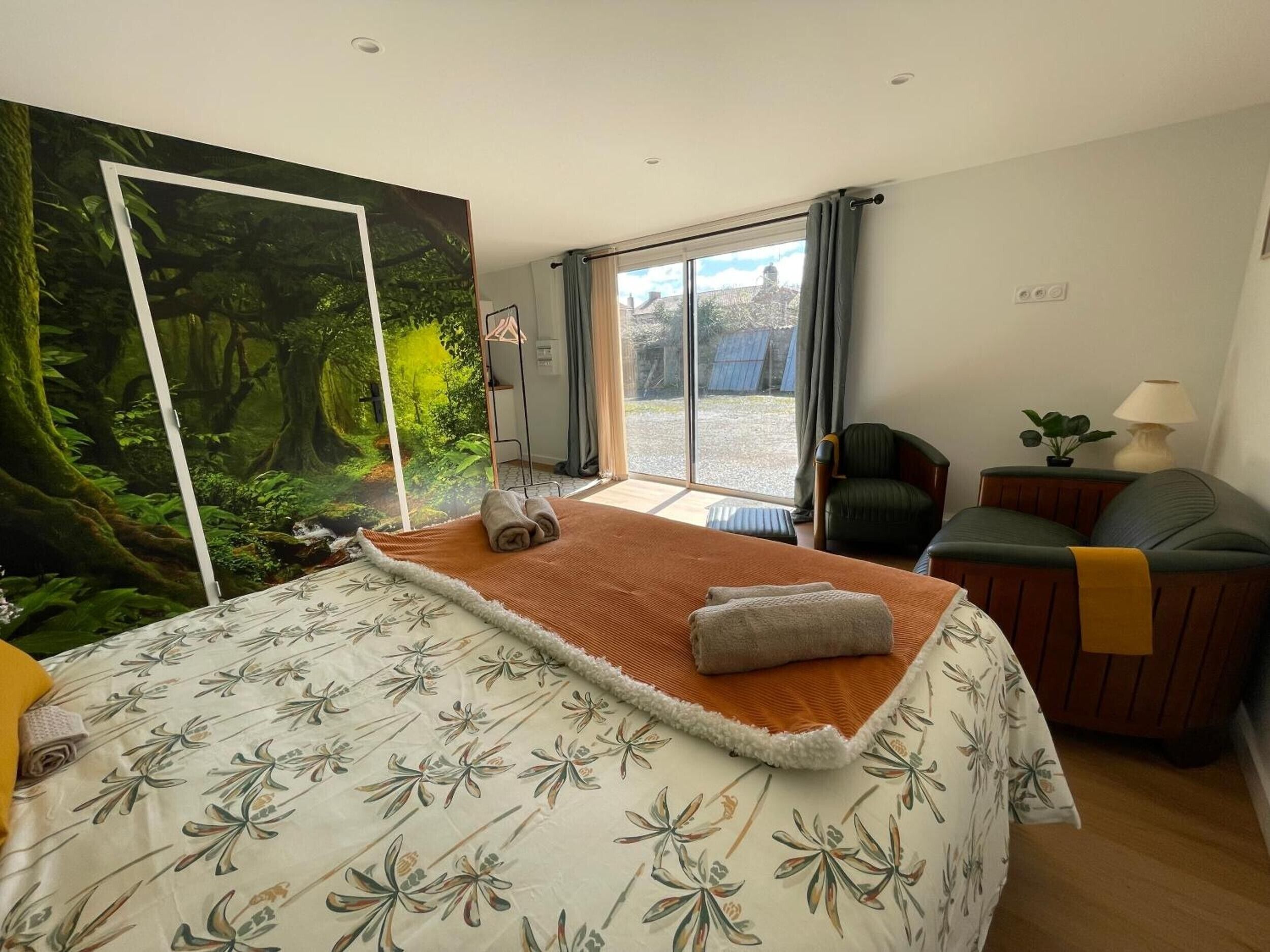 Une chambre avec un grand lit, une vue sur une forêt par une porte coulissante, un fauteuil, une lampe et une petite table.