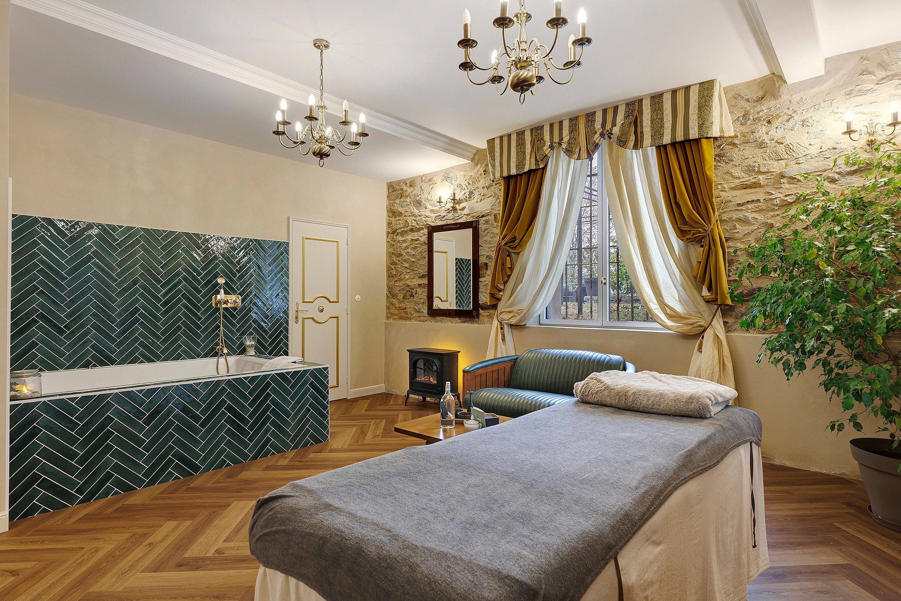 A room with a stone wall, a wooden floor, a bed, a bench, a small table, a mirror, and a chandelier.