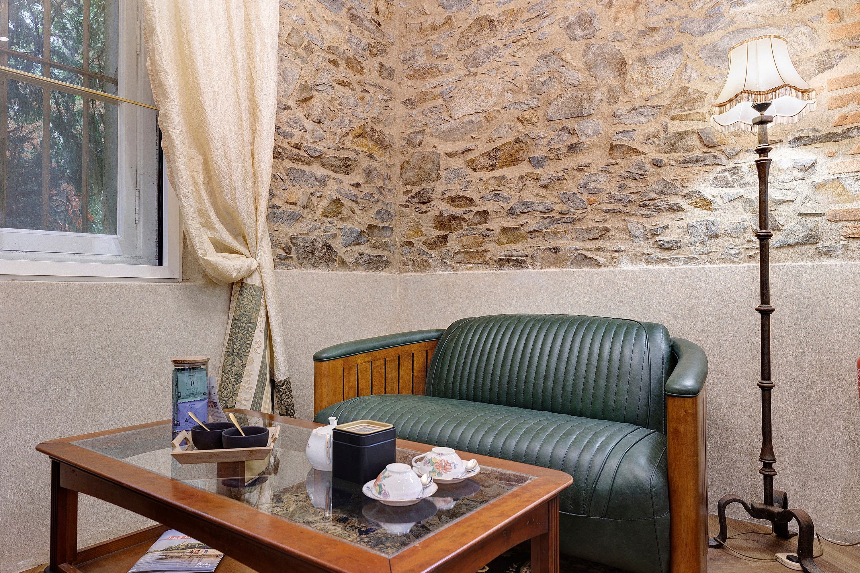 A room with a stone wall, a green leather sofa, a wooden table with a glass top, and a floor lamp.