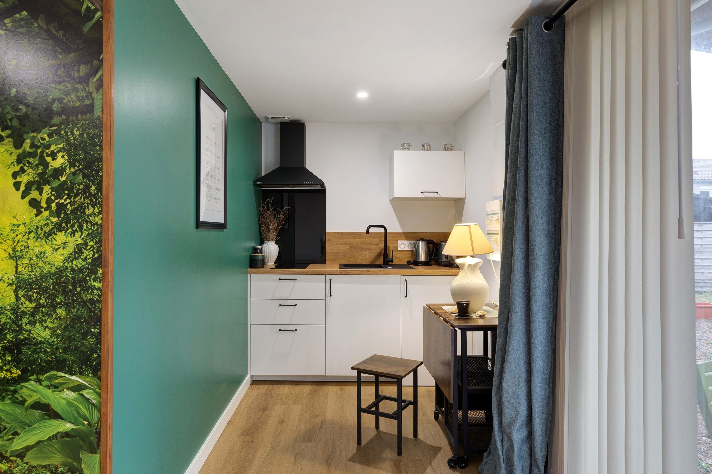 Una cocina compacta con armarios blancos, extractor de aire negro, encimera de madera y una pequeña mesa con lámpara.