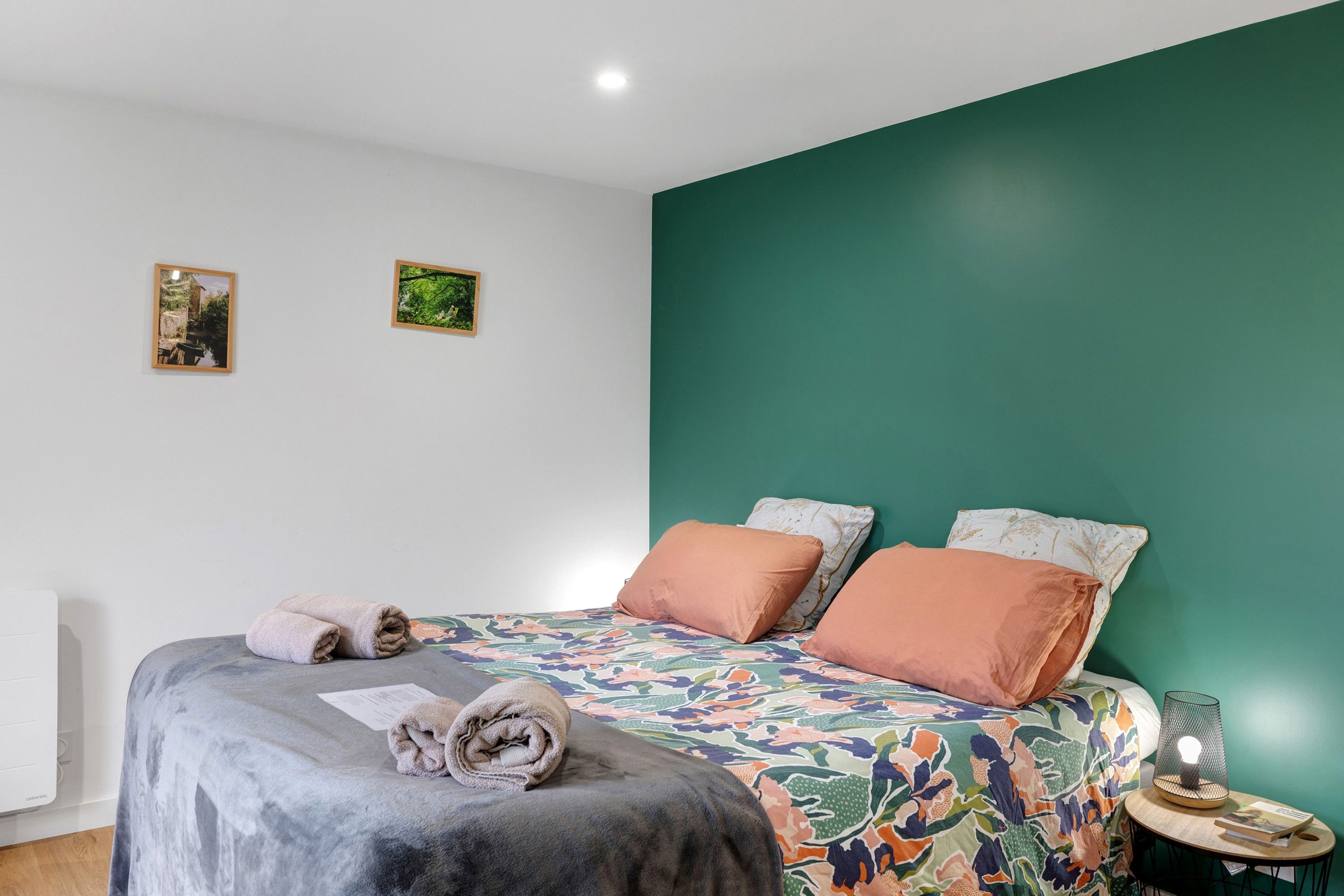 A neatly made bed with a floral bedspread, two pillows, and two rolled towels on a white wall.