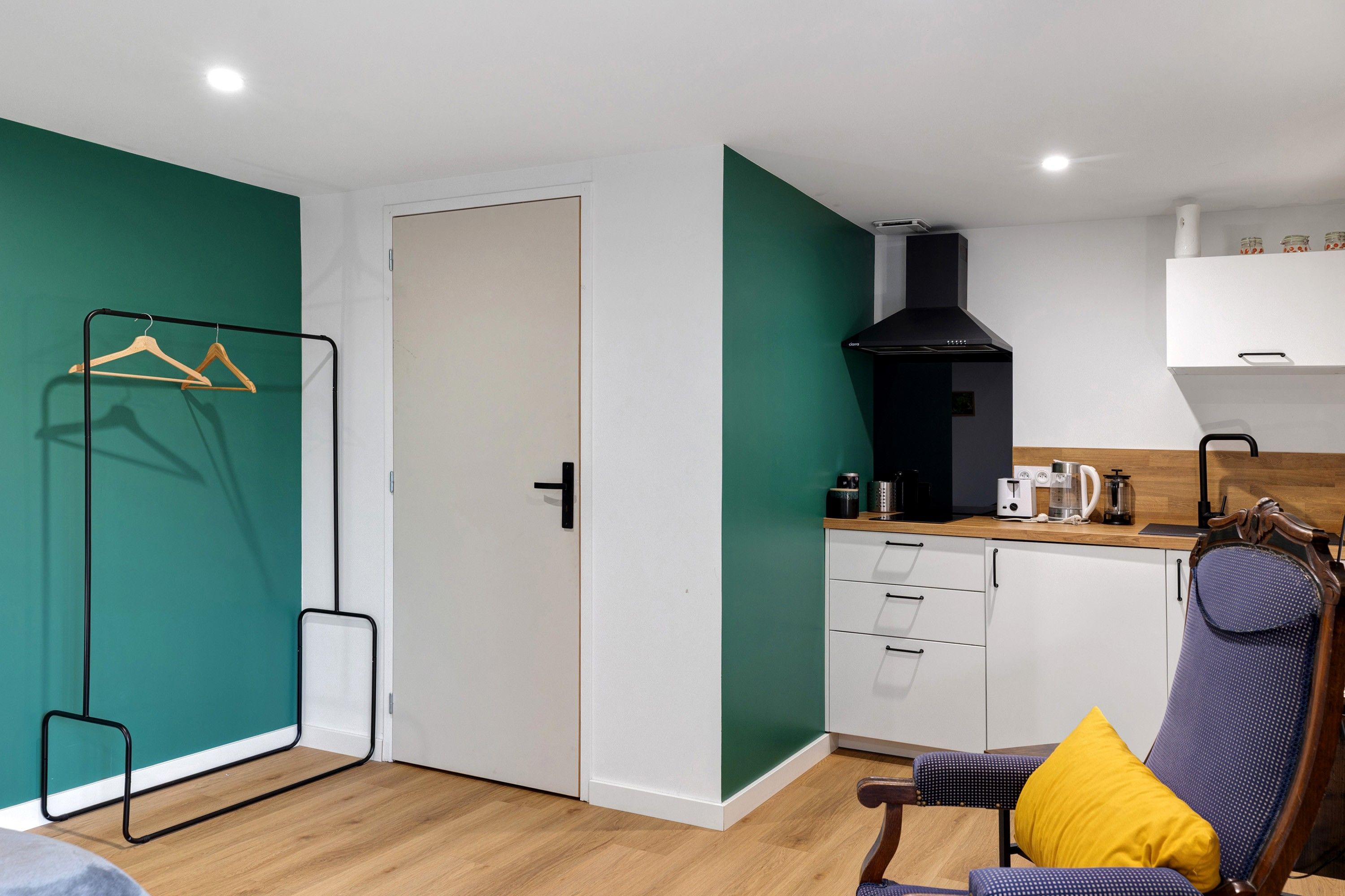 A compact living space with a kitchenette, a wooden chair with a yellow cushion, and a coat rack.