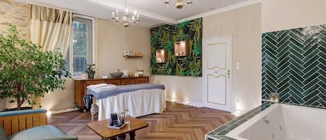 Una habitación bien iluminada con piso de madera, un ventanal con cortinas, un candelabro, una camilla de masaje, un banco de madera, una mesa pequeña con objetos, y una pared verde con patrón de espiga.
