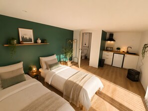 Een slaapkamer met twee bedden, een nachtkastje met een lamp, een hangplant en een ingelijst beeld aan de muur.