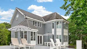 Ein zweistöckiges Haus mit überdachter Terrasse, die Sitzgelegenheiten und Sonnenschirme bietet.