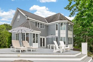 Ein zweistöckiges Haus mit überdachter Terrasse, die Sitzgelegenheiten und Sonnenschirme bietet.