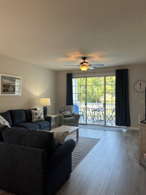 A living room with a sectional sofa, a patterned armchair, a ceiling fan, and a view through a glass door to an outdoor patio. - Golf and Beach Condo!! Sleeps 8! Labor Day Special! Resort amenities! (North Myrtle Beach)