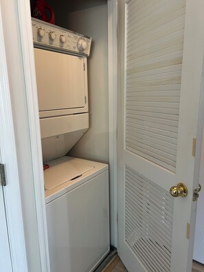A compact laundry room with a stacked washer and dryer, a small shelf, and a door with a louvered design. - Golf and Beach Condo!! Sleeps 8! Labor Day Special! Resort amenities! (North Myrtle Beach)