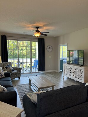 A living room with a sofa, coffee table, and a TV on a cabinet. There is a ceiling fan, a clock, and a view of the outdoors through a sliding glass door. - Golf and Beach Condo!! Sleeps 8! Labor Day Special! Resort amenities! (North Myrtle Beach)