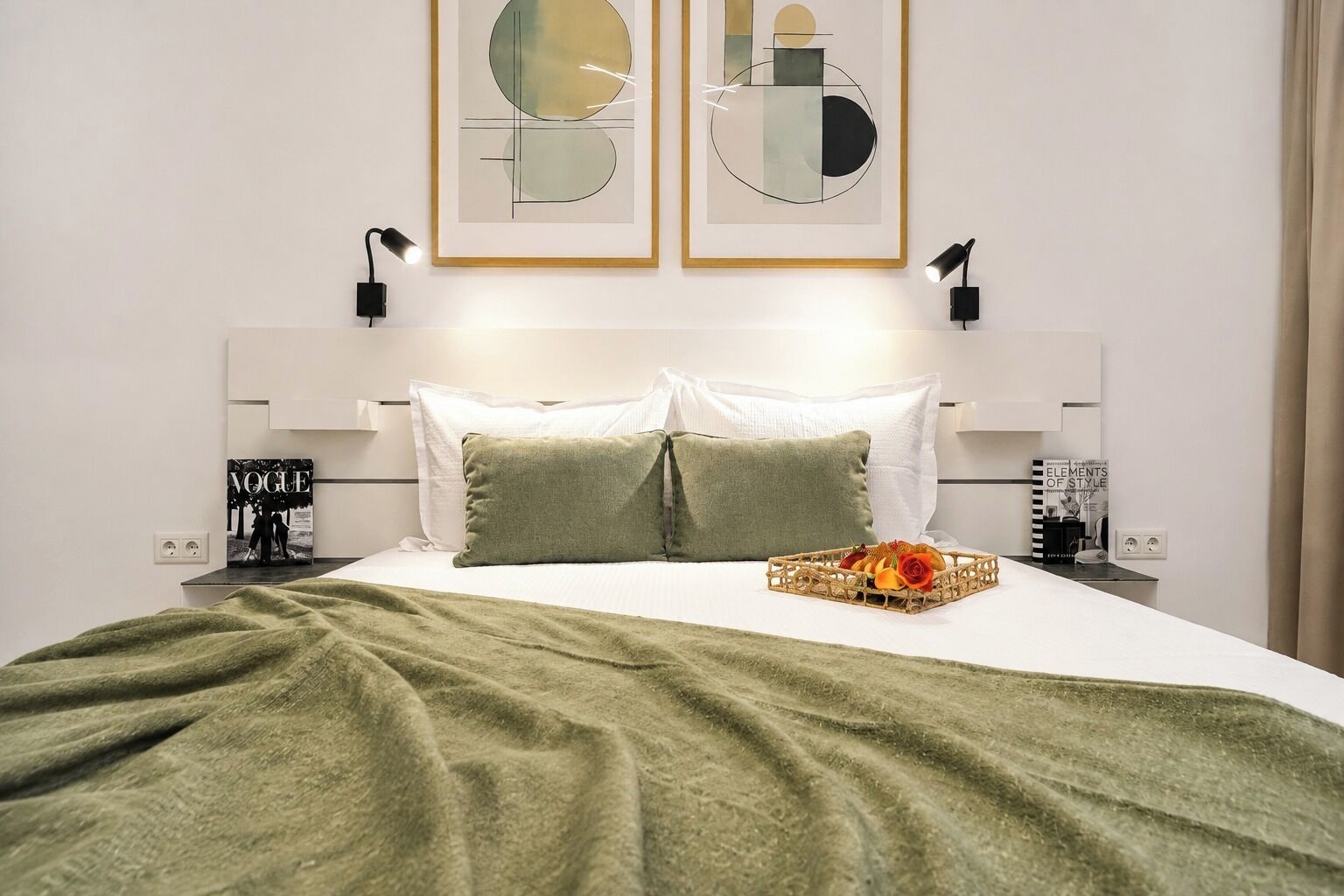 Un llit ben arreglat amb una manta verda, dos almoiners i un safata decorativa amb flors. Hi ha dues làmpades penjades a la paret i dues obres d'art abstractes emmarcades sobre el llit.
