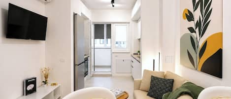 A modern living room with a sofa, a flat-screen TV, and a kitchen in the background.