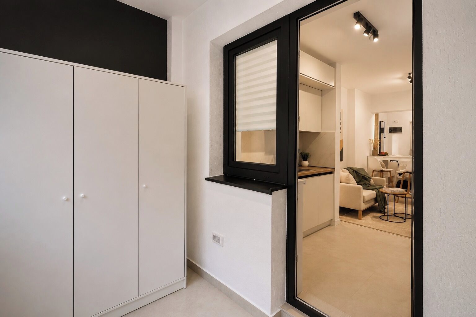 A modern interior with a wardrobe, a window with blinds, and a glimpse of a living area with a sofa and a table.