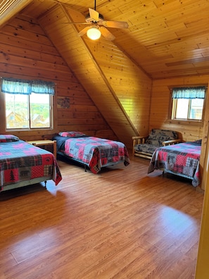 A cozy attic bedroom with three beds, wooden floors, and a sloped ceiling with a ceiling fan.