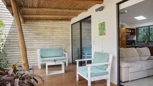 A wooden deck with two white chairs and a table, a sliding glass door leading to a kitchen, and a white sofa in the background. - Eco Casita 1.8 in Hacienda Iguana (Tola)