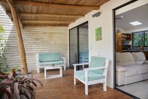A wooden deck with two white chairs and a table, a sliding glass door leading to a kitchen, and a white sofa in the background. - Eco Casita 1.8 in Hacienda Iguana (Tola)