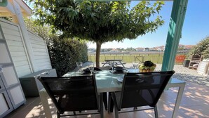 Une terrasse extérieure avec une table dressée pour deux personnes, entourée de chaises, d’un verre d’eau et d’un panier de fruits.