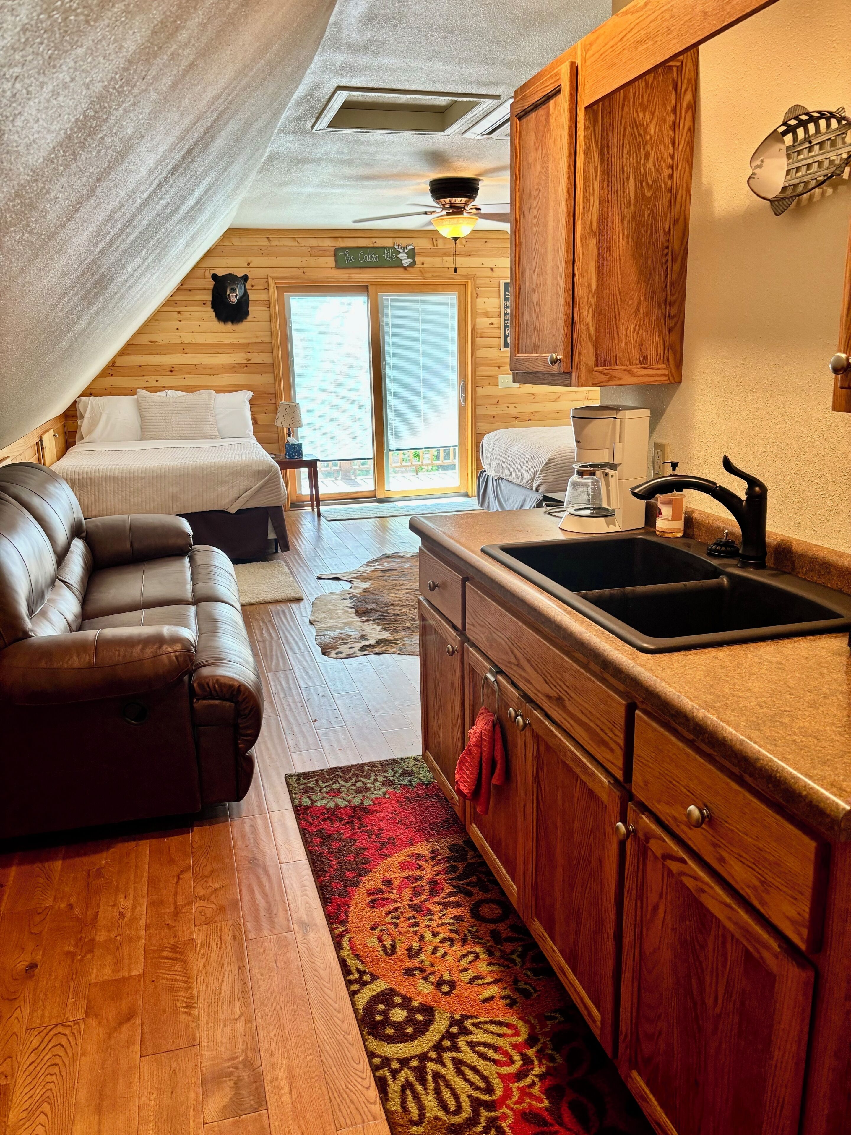 A cozy cabin interior with a bed, a sofa, a kitchen area, and a sliding glass door leading to a deck.