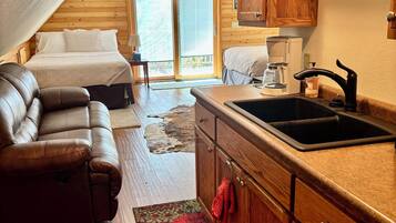 A cozy cabin interior with a bed, a sofa, a kitchen area, and a sliding glass door leading to a deck.