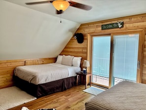 Et hyggeligt soveværelse med skrå vægge, en seng med brun dynebetræk, et natbord med en lampe, og et skilt med teksten "The Cabin Life".