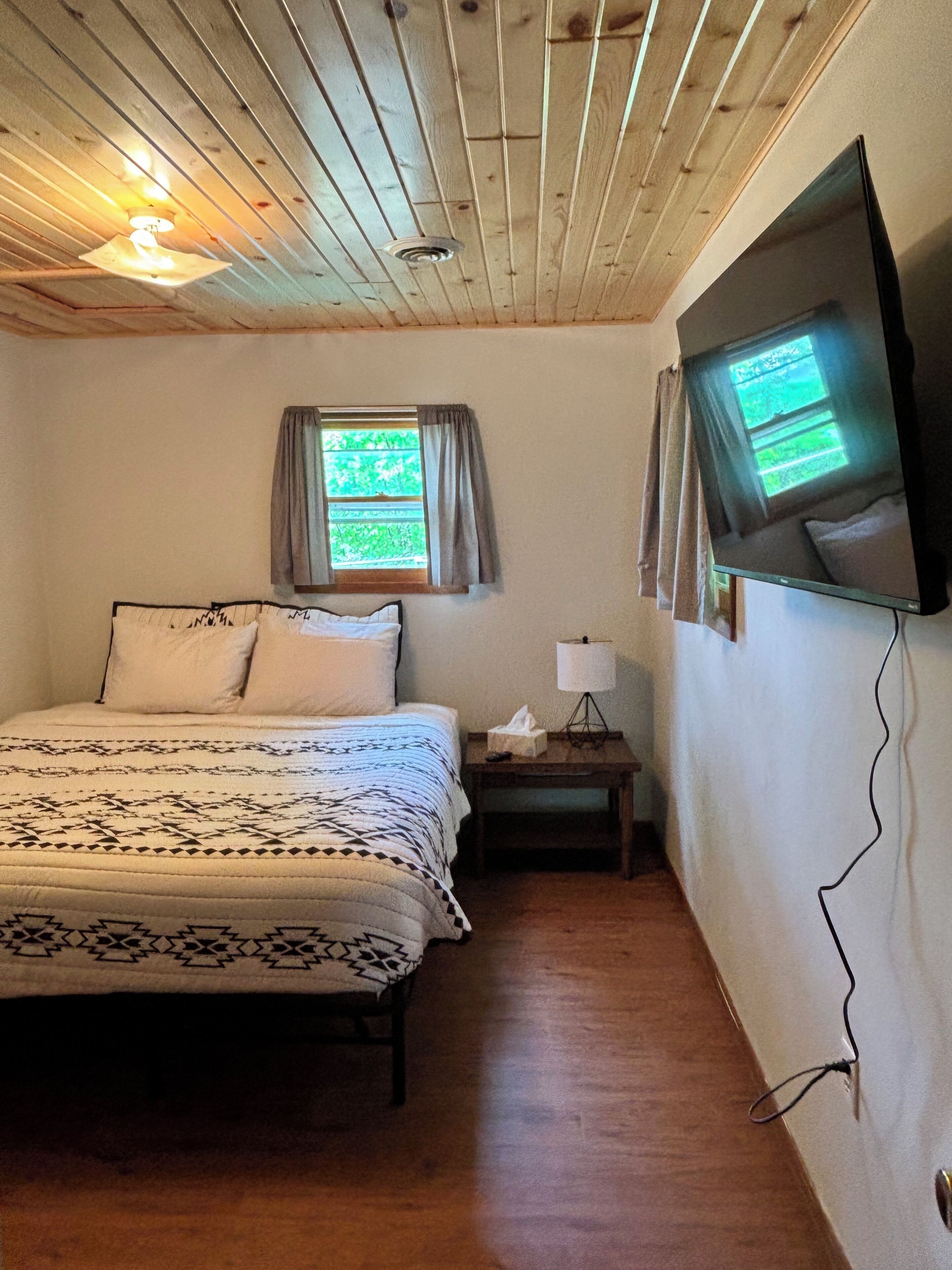 Une chambre à coucher avec un lit, une fenêtre avec des rideaux, une table de chevet avec une lampe et un téléviseur fixé au mur.