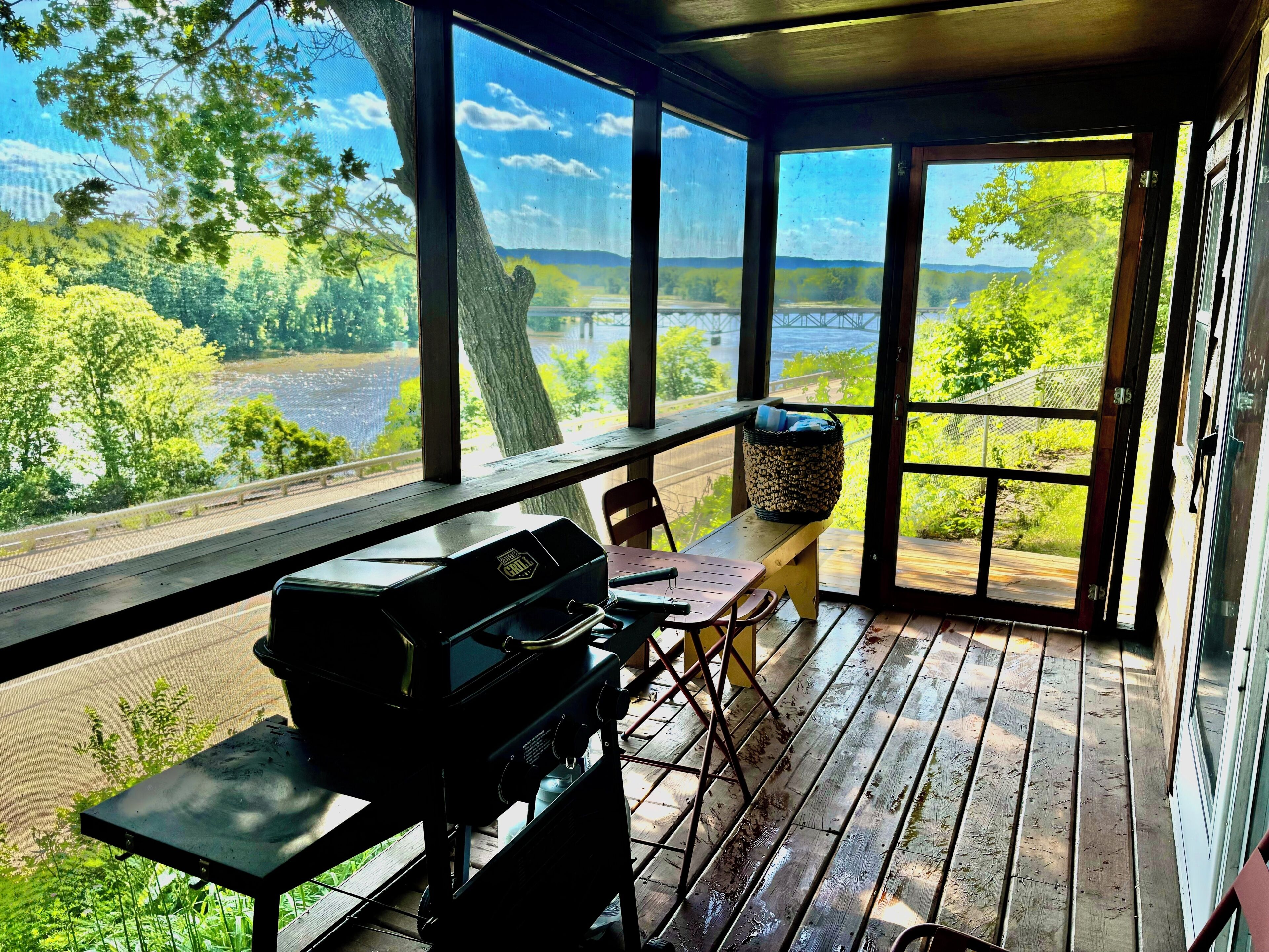 Ein überdachter Außenbereich mit Grill, Tisch und Bank mit Blick auf einen Fluss und viel Grün.