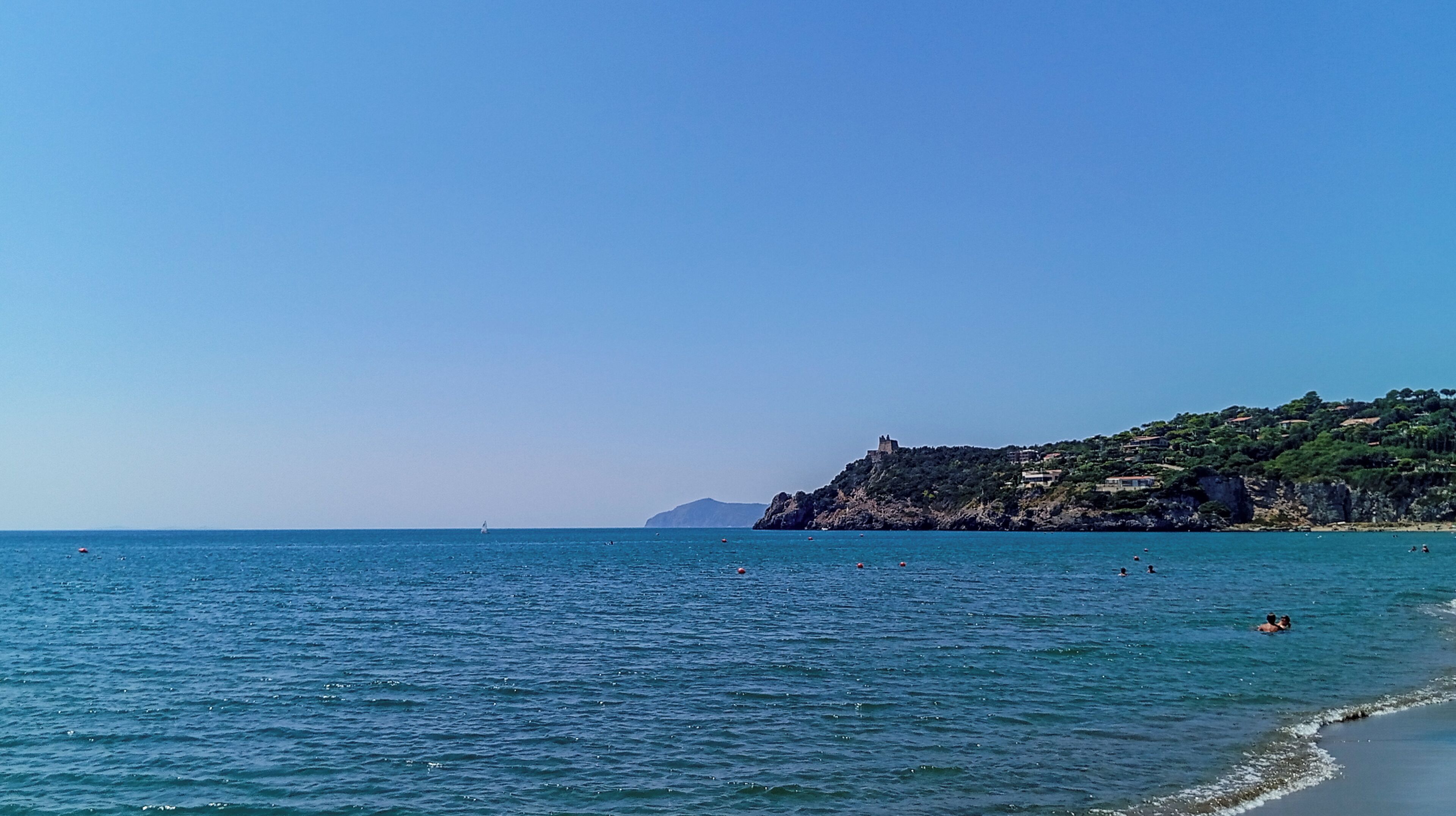 A coastal landscape with a clear sky, calm sea, and a rocky hill with buildings.