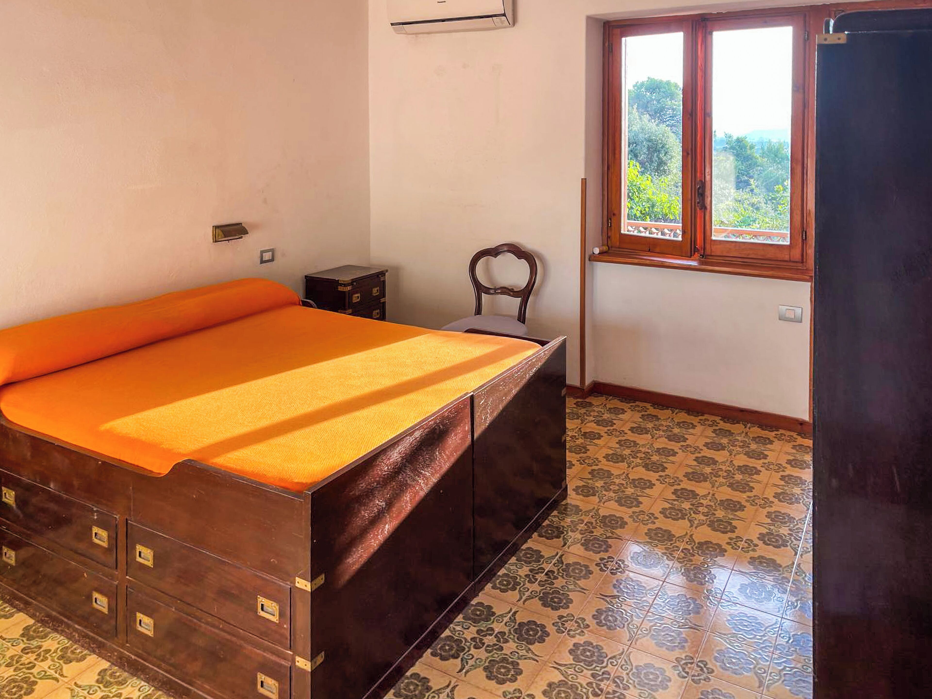 Une chambre à coucher avec un lit, une armoire en bois, une chaise, une fenêtre et une porte.