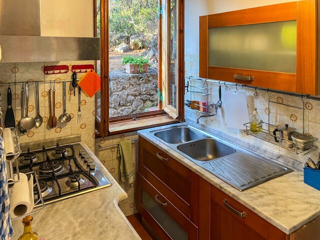 Una cocina con estufa a gas, pileta doble, gabinetes de madera y ventana con vista a follaje.