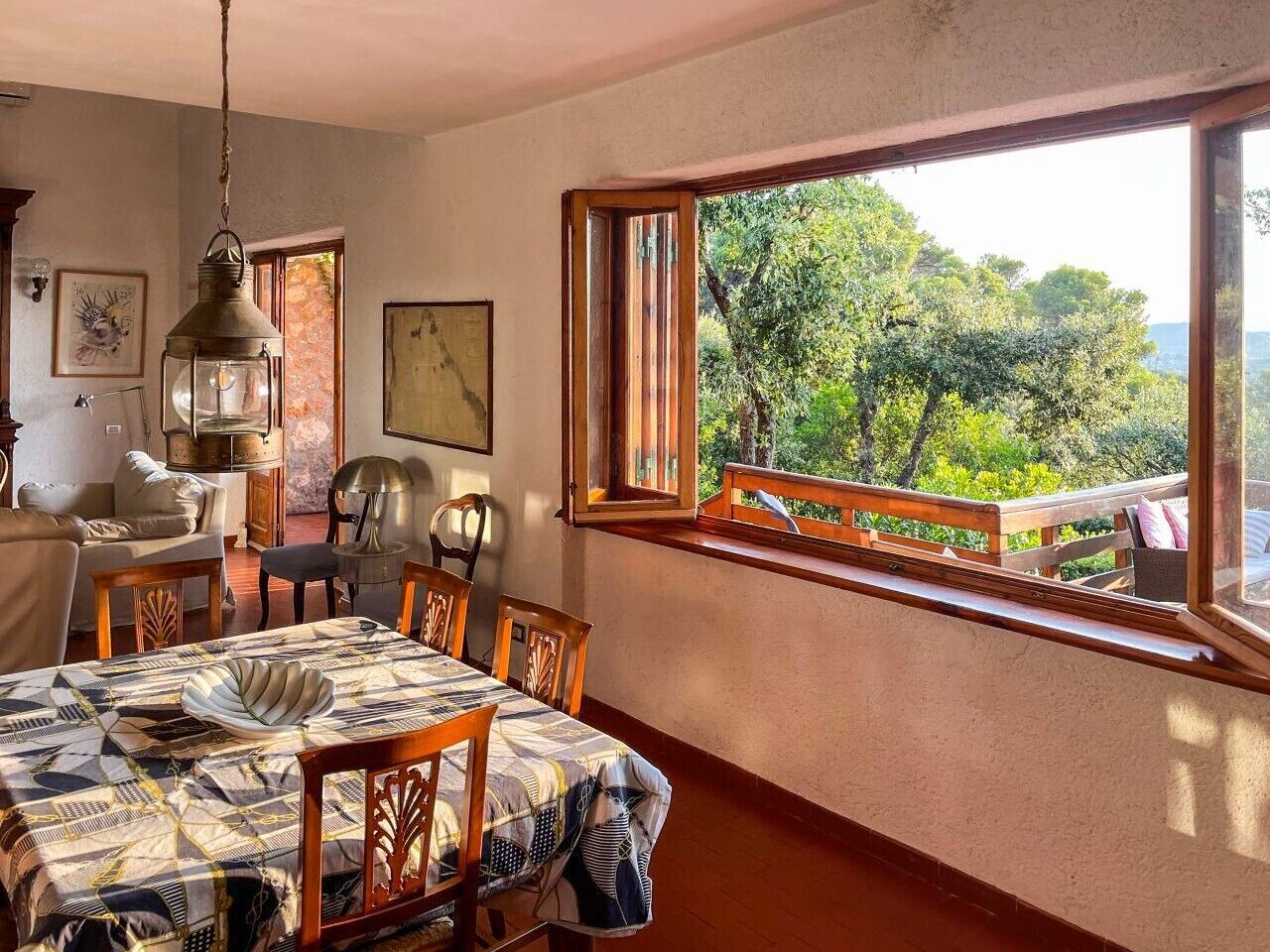 Uma área de refeições com uma mesa posta para quatro pessoas, um candeeiro pendente e uma janela com vista para o verde.