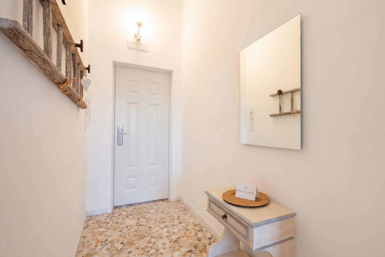 Un corridoio con una porta bianca, un appendiabiti in legno, uno specchio e una piccola toeletta di legno con una targhetta.