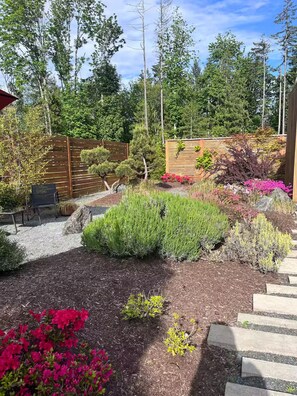 A landscaped garden with a variety of plants and shrubs, a wooden fence, and a stone pathway. - Sustainable Garden Suite Retreat - Modern, serene & peaceful oasis (Nanaimo)