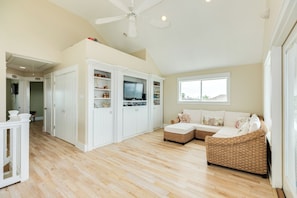 A spacious living room with a high ceiling, a built-in entertainment unit, a large window, and a wicker sofa. - Pet Friendly Canal Home w/ Top Level Viewing Deck,  Boat Slip & More! (Galveston)