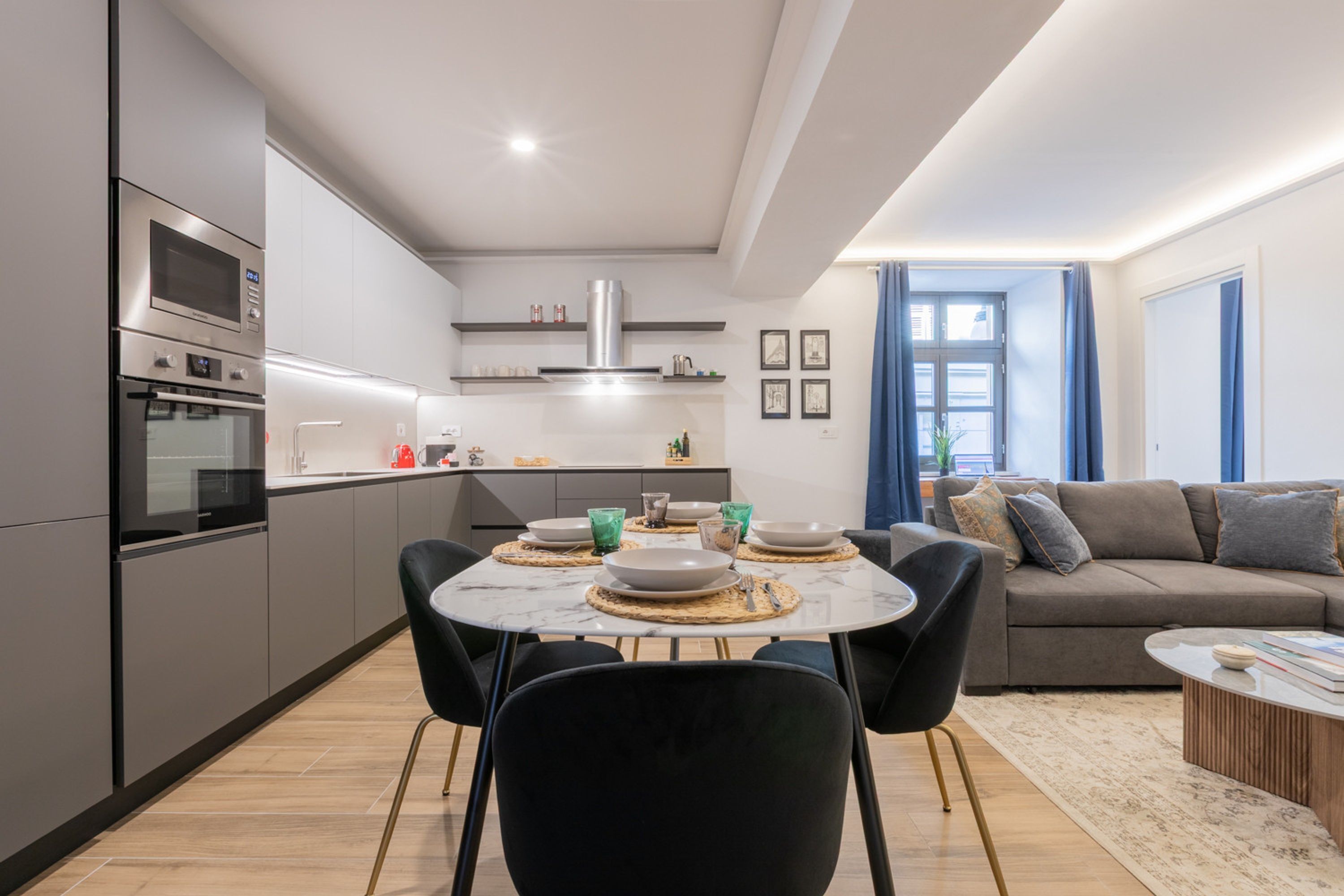 Una cuina moderna amb electrodomèstics integrats, una zona de menjador amb una taula rodona i cadires, i un saló amb un sofà i una taula de centre.