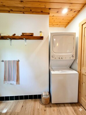 Een wasruimte met een wasmachine en droger, een plank met een pot en een bord, een handdoekhouder met een kleurrijke handdoek, en een mand op de vloer.