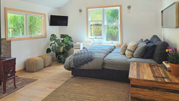 A bedroom with a sloped ceiling, a bed with a blanket, a TV, and two windows with views of greenery.