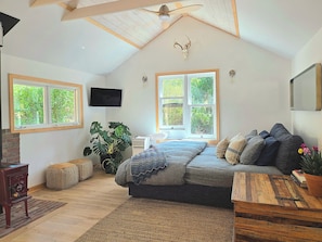 Ein Schlafzimmer mit schrägen Wänden, einem Bett mit Decke, einem Fernseher und zwei Fenstern mit Blick auf Grünflächen.