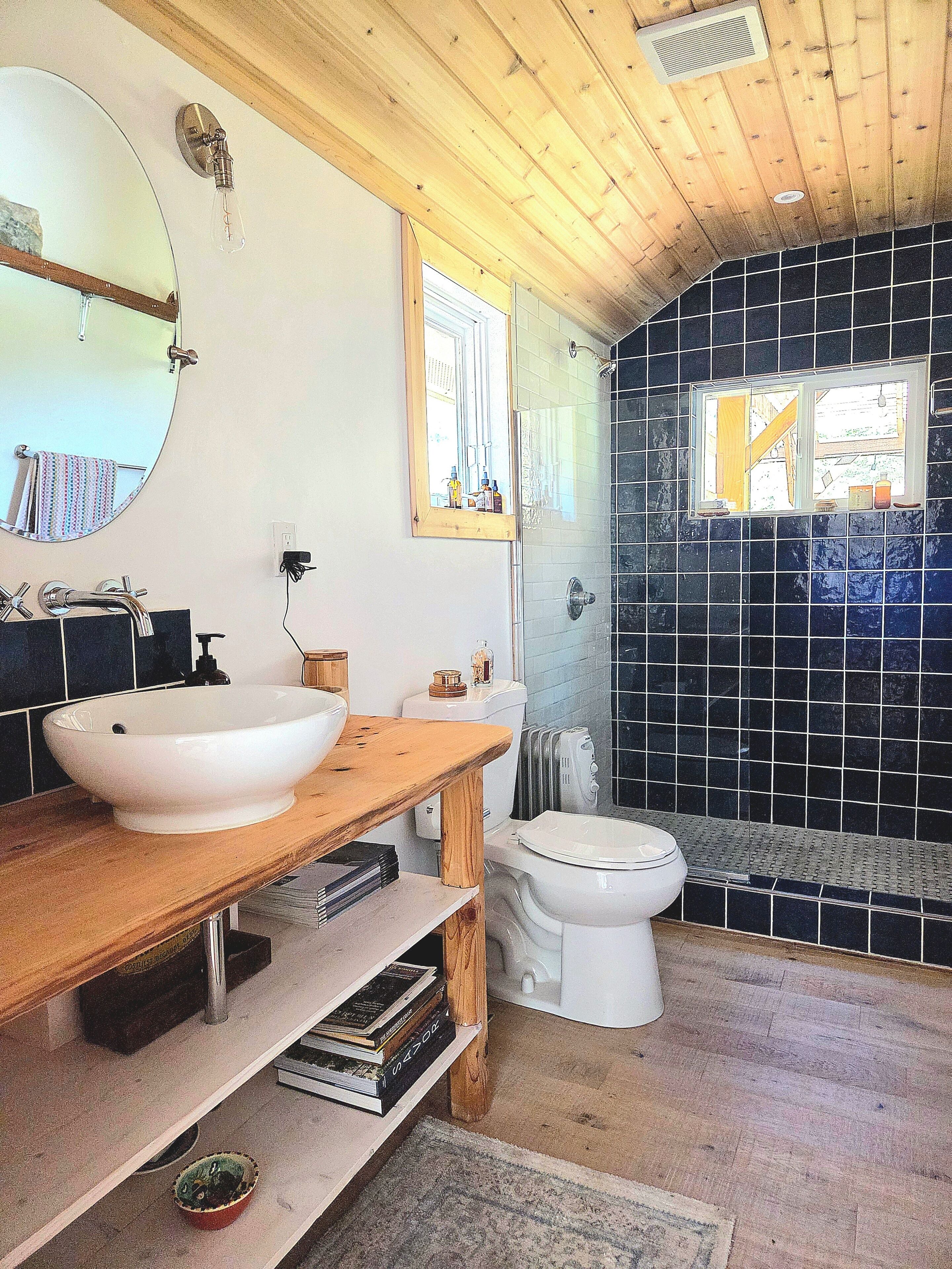 A bathroom with a wooden vanity, a round mirror, a white sink, a toilet, and a shower area with black tiles.