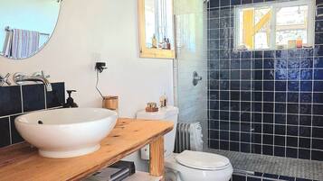 A bathroom with a wooden vanity, a round mirror, a white sink, a toilet, and a shower area with black tiles.