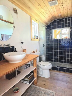 A bathroom with a wooden vanity, a round mirror, a white sink, a toilet, and a shower area with black tiles.