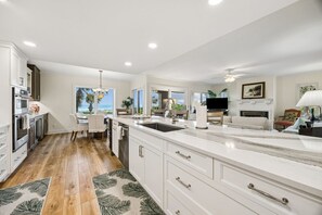 A modern kitchen with white cabinetry, a large island with a sink, and a dining area with a view of the ocean. - 1827 - Sanctuary On Amelia Island (Fernandina Beach)