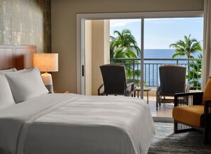 Habitación de hotel con una cama grande, una mesita de noche con lámpara y vistas al océano a través de una puerta corrediza de vidrio.