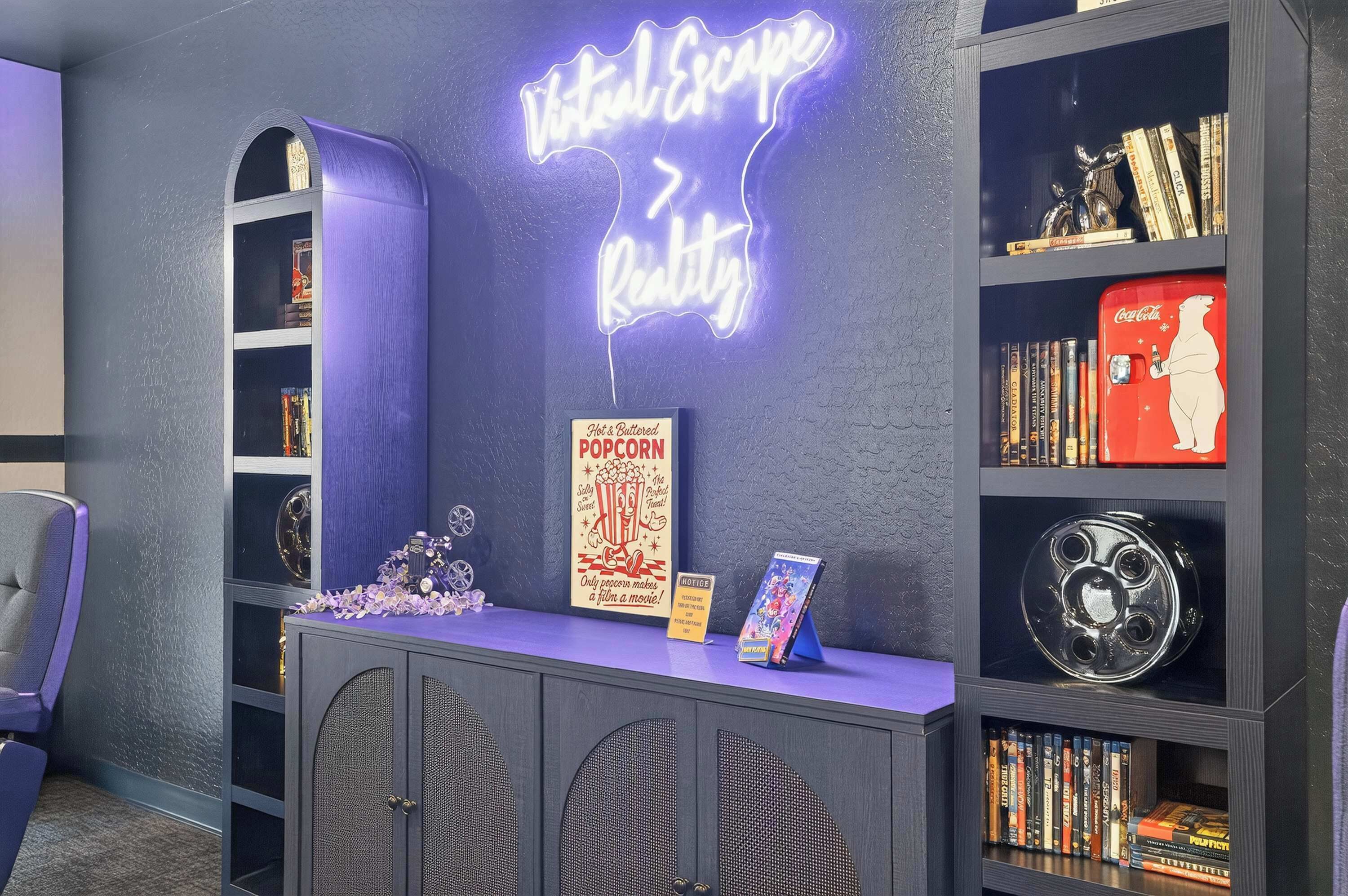 Ein Zimmer mit einem Neonlichtschild mit dem Schriftzug 'Virtual Escape Reality', einem Schild mit der Aufschrift 'Popcorn' sowie einer Sammlung von Büchern und DVDs in Regalen.