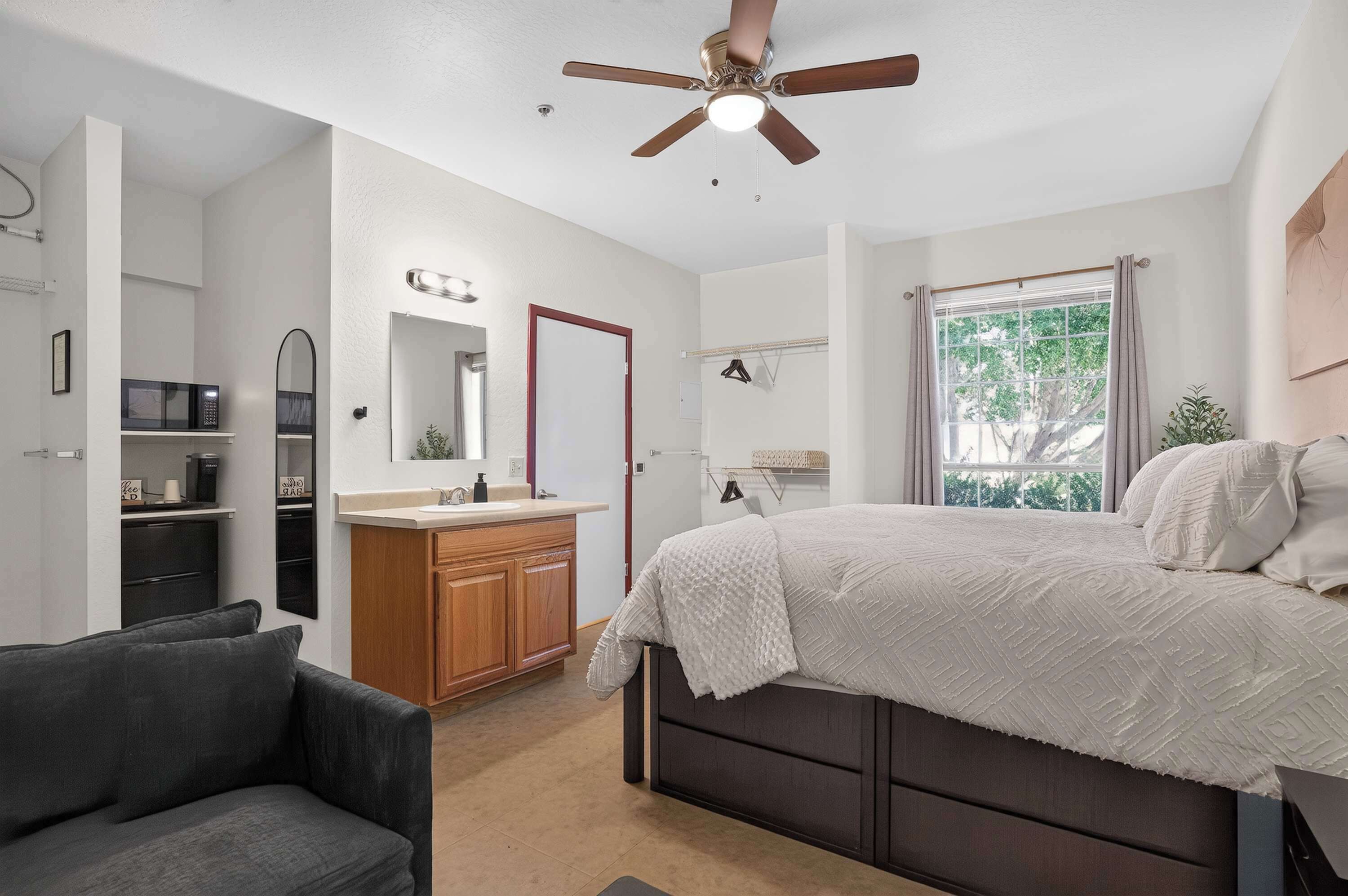 A bedroom with a bed, a ceiling fan, a window with curtains, a bedside table, and a chair.