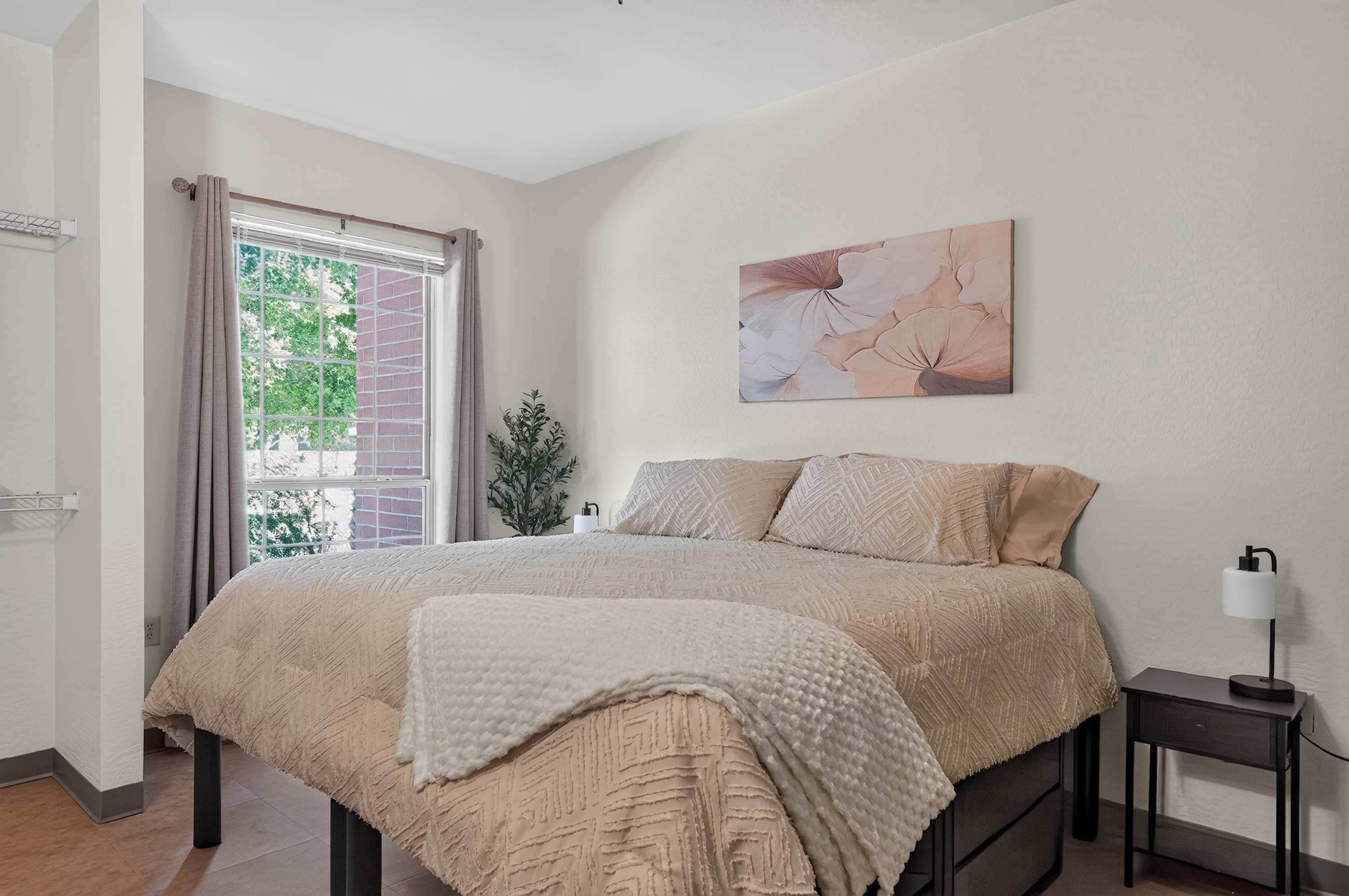 Un dormitorio con cama, mesita de noche, lámpara, ventana con cortinas y un cuadro colgado en la pared.
