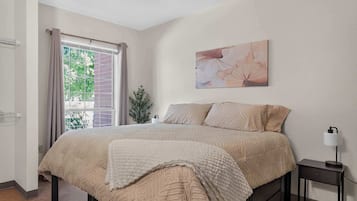 A bedroom with a bed, a nightstand, a lamp, a window with curtains, and a wall-mounted painting.