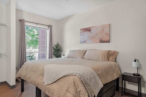 A bedroom with a bed, a nightstand, a lamp, a window with curtains, and a wall-mounted painting. - Modern King Suite - Steps from UAT and near ASU (Tempe)