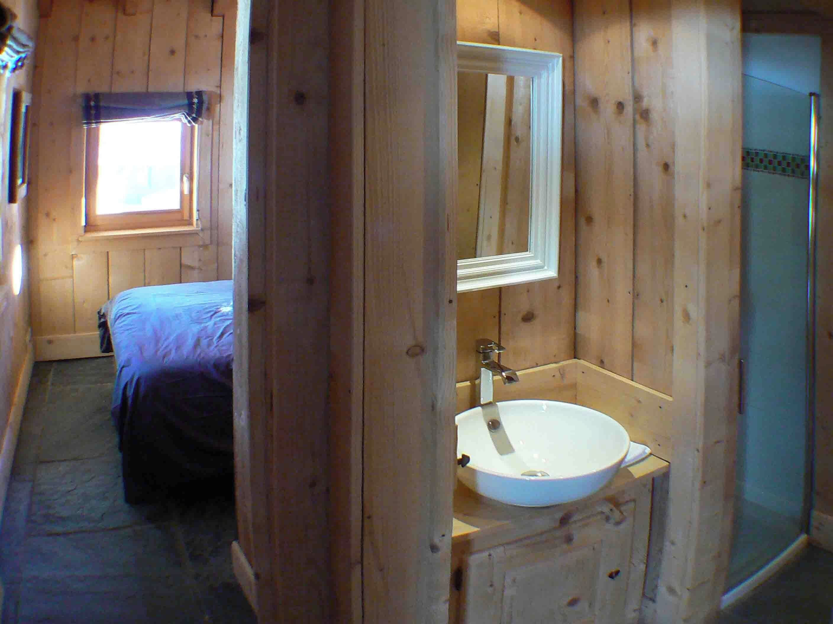 Une chambre dans un chalet en bois, avec un lit, une salle de bain comprenant un lavabo et un miroir, et une fenêtre.