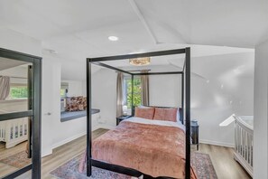 A modern bedroom with a four-poster bed, a small bedside table, a window with curtains, and a view of greenery outside.