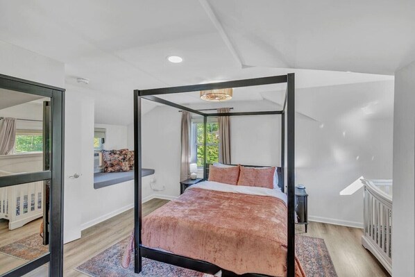 A modern bedroom with a four-poster bed, a small bedside table, a window with curtains, and a view of greenery outside.