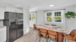 A modern kitchen with a stainless steel refrigerator, a marble dining table with six chairs, and a window with a view of greenery. - Family Retreat with Pool & Playground (Roslyn)